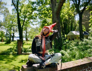 Artist in residence Alex Tatarsky, wearing a gnome costume, looks up towards the sky. Trees and grass are visible in the background.