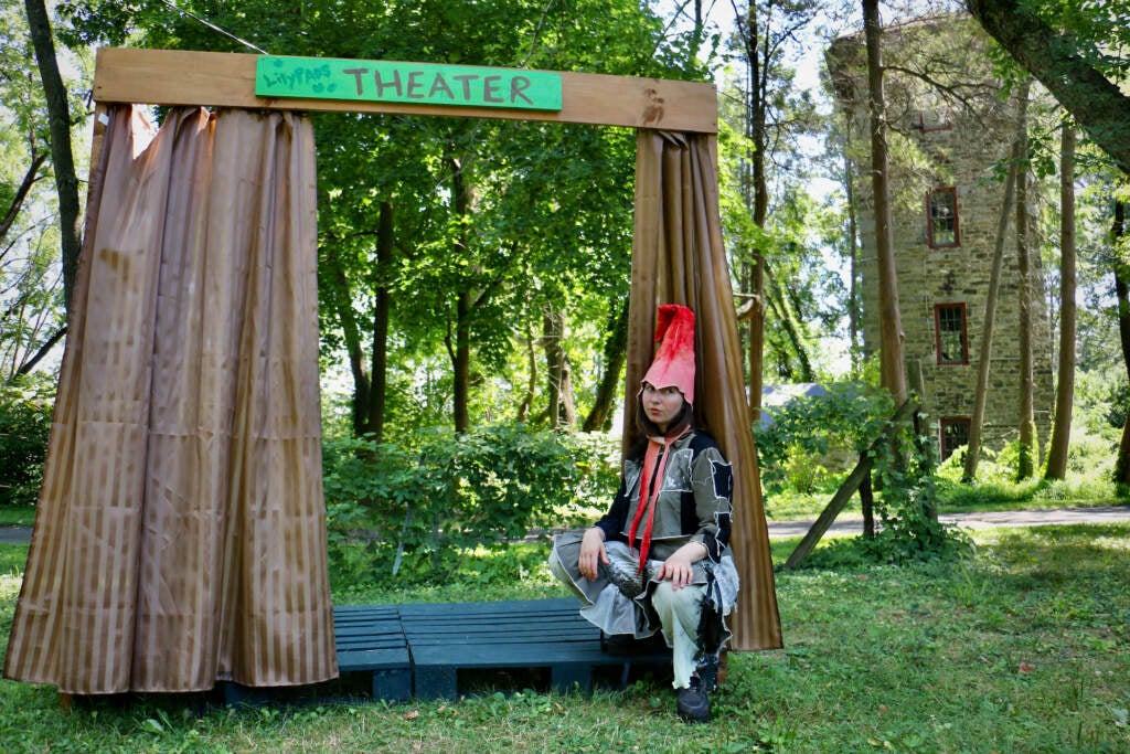 Alex Tatarsky, in a garden hermit costume, sits next to a small, homemade theater stage.