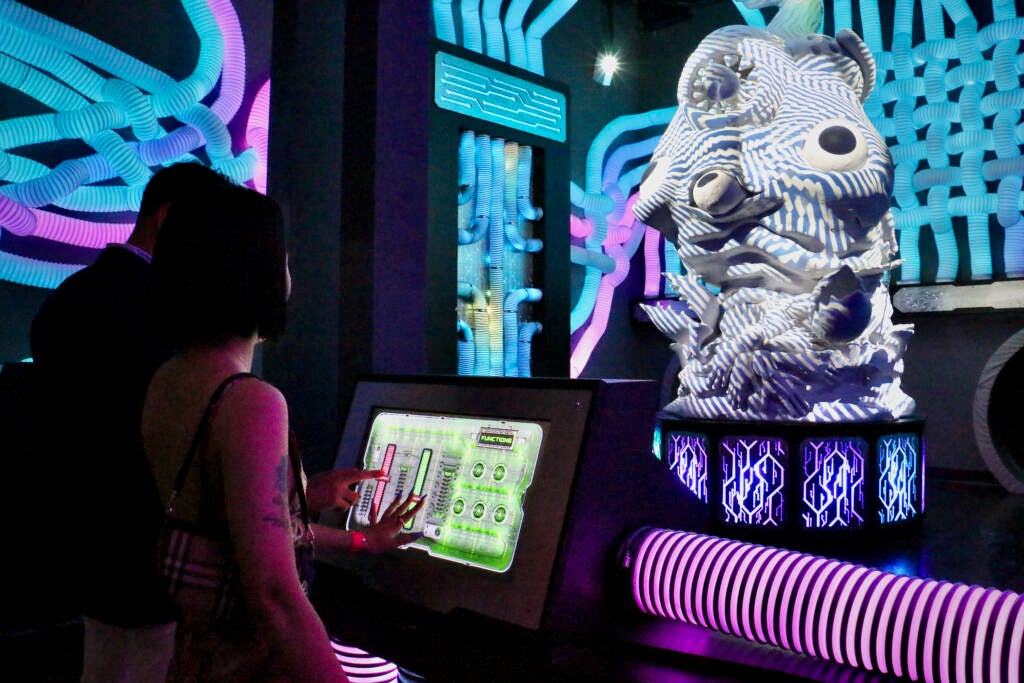 People press buttons on a touch screen. A strange sculpture is in front of them and neon-lighted pipes criss-cross in the background.