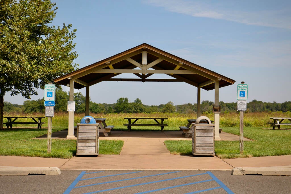 A gazebo stands next to a parking lot. In the distance, a meadow and trees are visible.