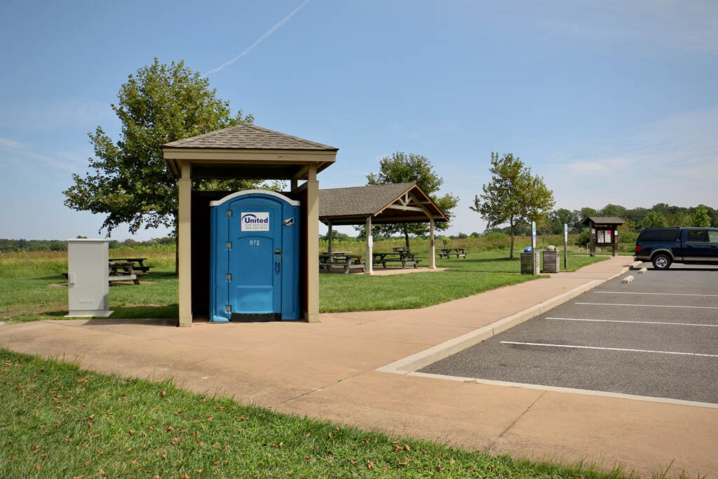 An outdoor port-a-pottie is visible at the edge of a meadow and a parking lot.