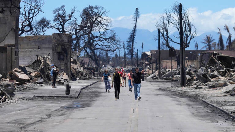 People walk down a road with damaged and burned wreckage visible on either side