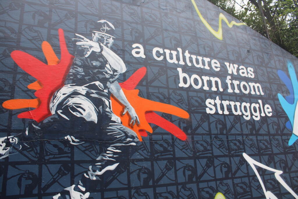 A b-boy is shown next to text that reads " a culture was born from struggle" on a mural