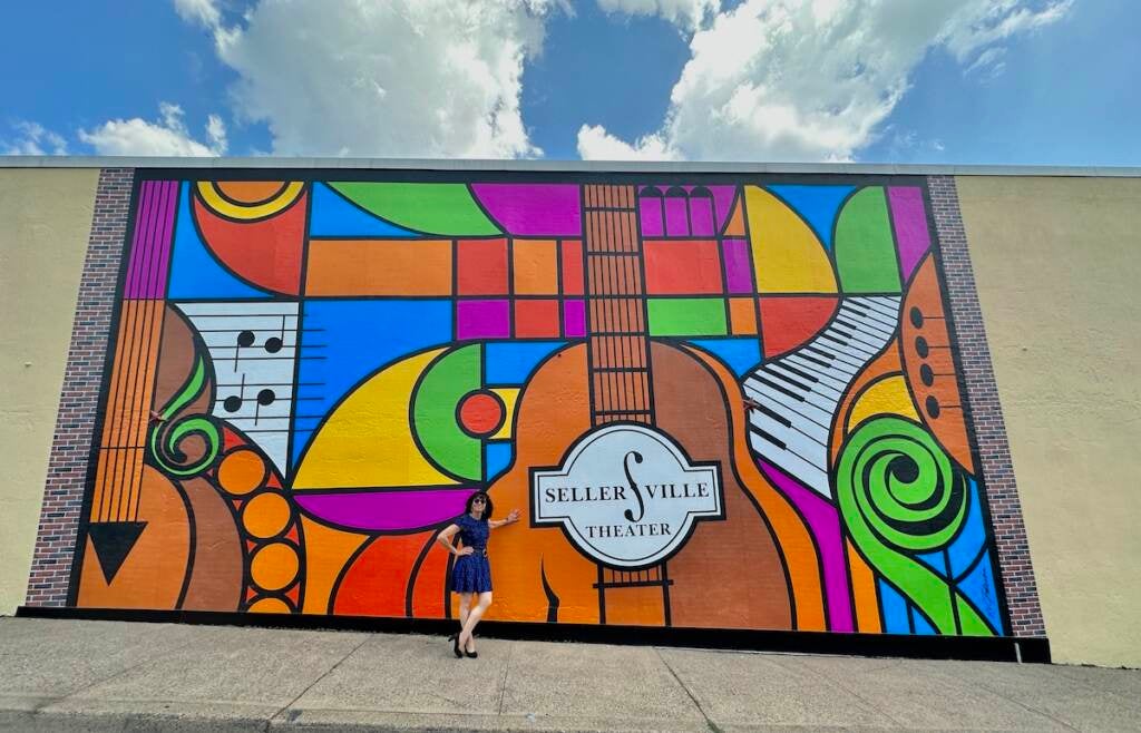 Elayne Brick poses in front of a mural with many colors that shows a guitar, keyboards, and the words "Sellersville Theater"