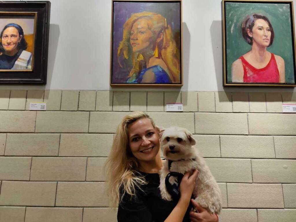 Anna Bogdanyk holding her dog, posing for a photo in front of her portrait.