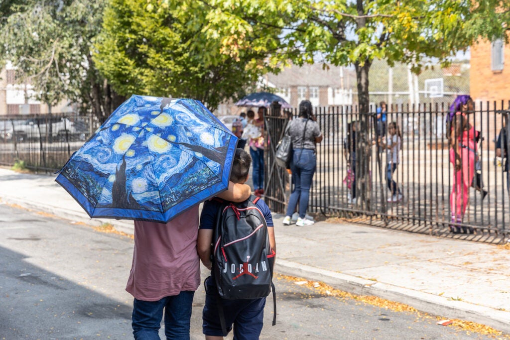 A person holding an umbrella walks with a student in the bright sun.