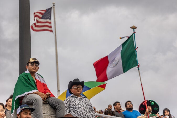 Two people watch on as members of a crowd. The U.S. flag and Mexican flag fly in the background.