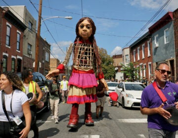 Little Amal, a 12-foot puppet, walks down a street in South Philadelphia, accompanied by residents and community members.
