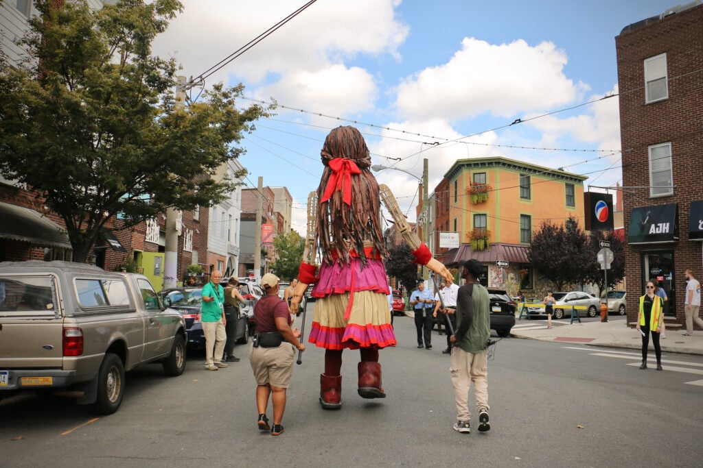 Little Amal walks down Passyunk Ave. with a group of people.