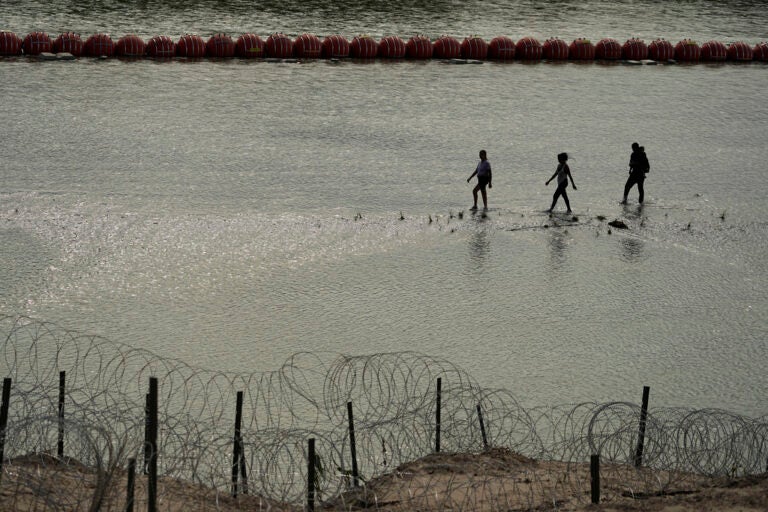 Migrants walk past large buoys being used as a floating border barrier on the Rio Grande, Monday, July 31, 2023, near Eagle Pass, Texas, as they cross from Mexico to the U.S. (AP Photo/Eric Gay)