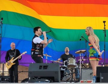 Kariselle Snow onstage as someone sings and a guitarist and drummer play in the background. In the back of the stage is a giant Pride flag.