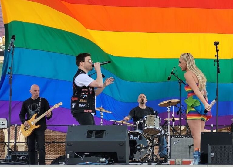 Kariselle Snow onstage as someone sings and a guitarist and drummer play in the background. In the back of the stage is a giant Pride flag.