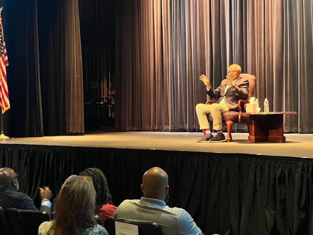 Dr. Clarence B. Jones speaks to an audience while seated onstage.