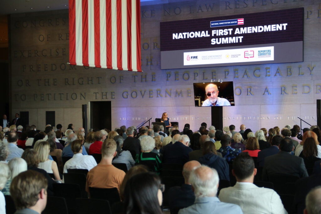 Salman Rushdie speaks from a screen to a large audience. A sign reads "National First Amendment Summit."