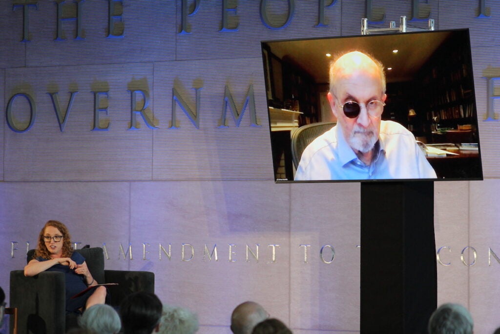 Suzanne Nossel speaks from a seat onstage. Salman Rushdie speaks from a screen overhead.