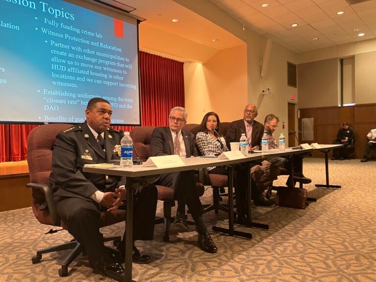 From left, Interim PPD Commissioner John Stanford, District Attorney of Philadelphia Larry Krasner and others participate in a panel, seated at a table, with a slideshow visible behind them.