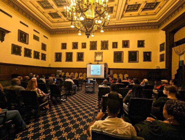 People seated in City Hall watch a documentary on the screen at the front of the room.