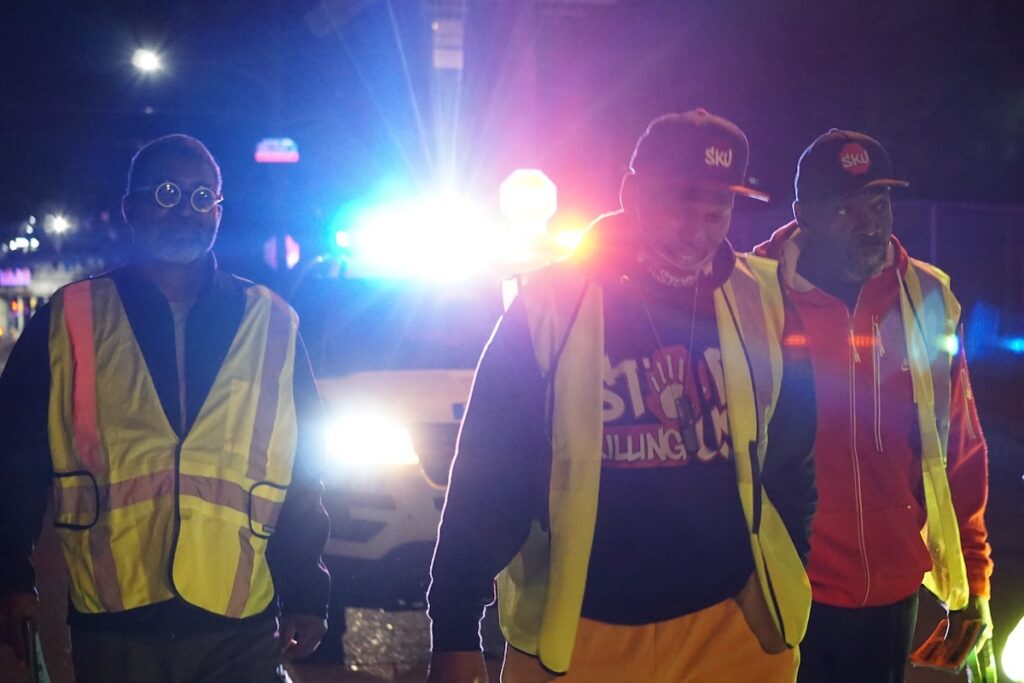 Jamal Johnson, center, with two other anti-violence activists walks at night wearing yellow safety vests.