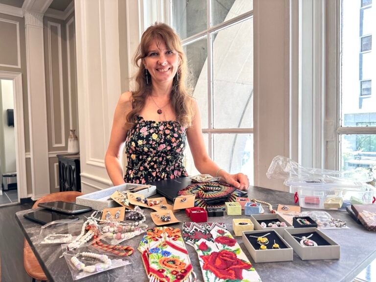 Oksana Pivush poses for a photo with her jewelry and other art displayed on a table in front of her.