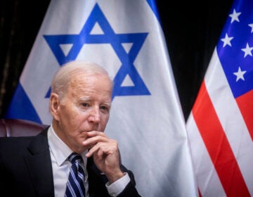 Biden sits in a chair, hand to face, with the Israeli and U.S. flags visible in the background.