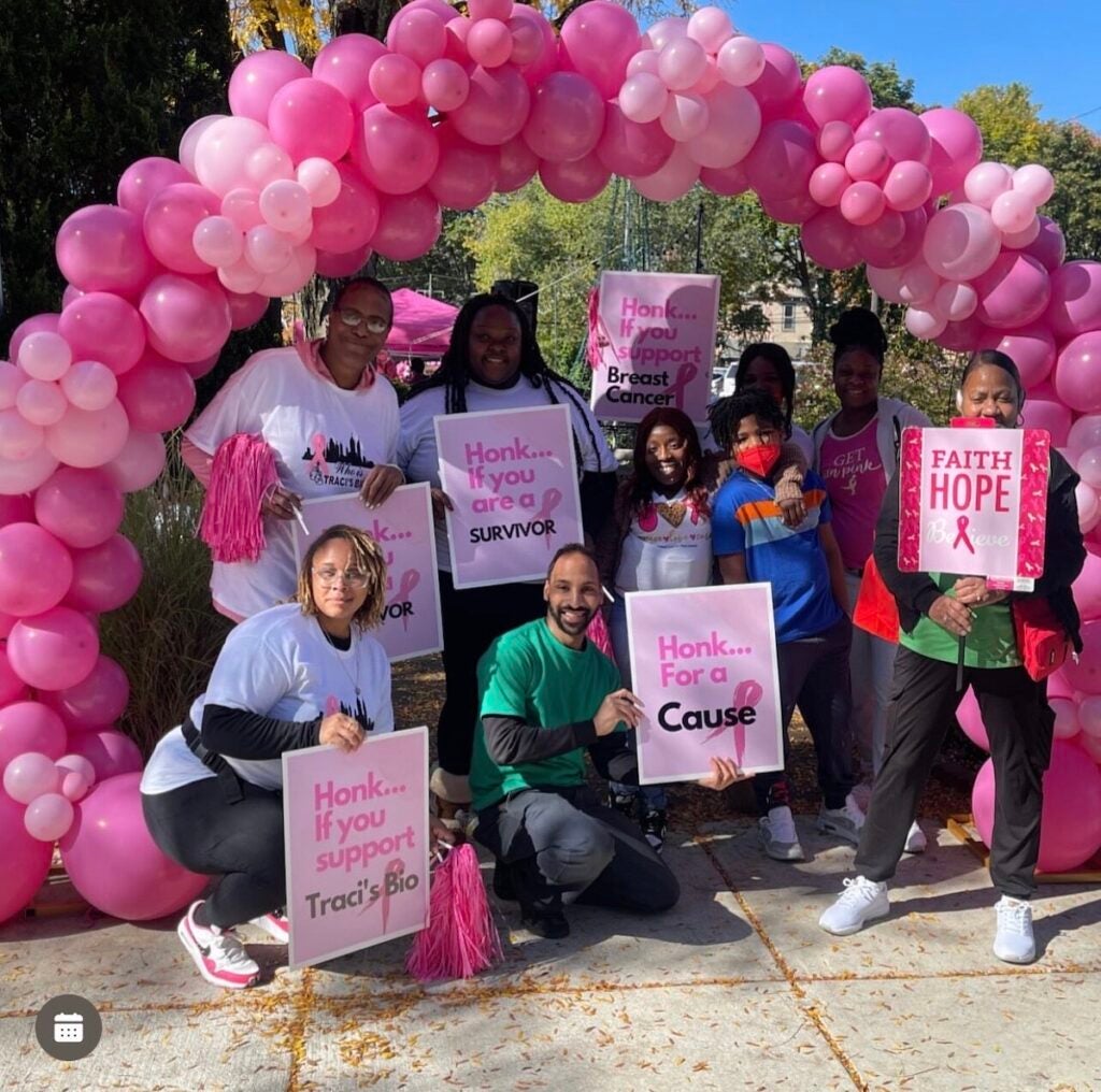 A group of people poses for a photo under an arbor of pink balloons holding signs reading Honk if you are a survivor.