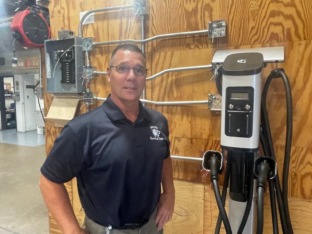 Bryan Bryant poses next to an EV charging station mounted on a wall in a workshop.