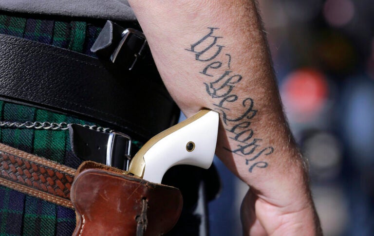 A close-up of someone's gun on a holster. On their arm is a tattoo that reads 