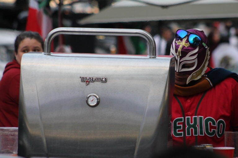 A person wearing a luchador mask and a Mexico jersey stands at a grill outside of the stadium.