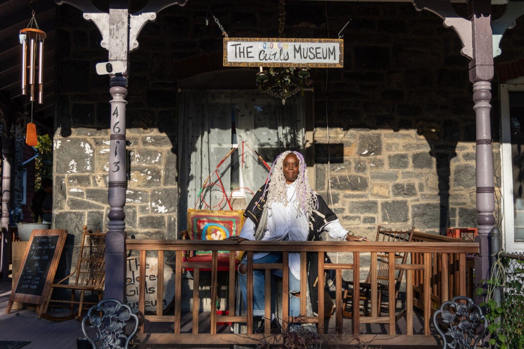 Vashti DuBois stands on her porch. Above her is a sign that reads The Colored Girls Museum.