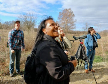 Nicole Seahorne Hameen looks upwards while holding a pair of binoculars. Other members of the bird watching group are visible in the background, also looking skyward.