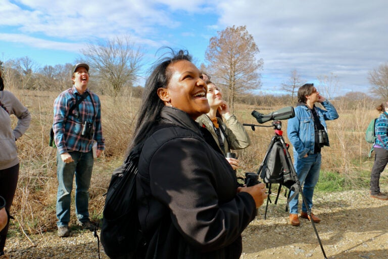 Nicole Seahorne Hameen looks upwards while holding a pair of binoculars. Other members of the bird watching group are visible in the background, also looking skyward.