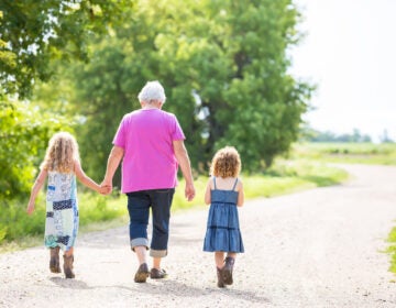 A grandmother walks down a road, holding the hands of two young children on either side of her.