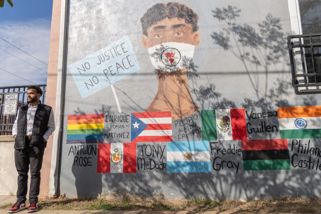 Branden Fletcher Dominguez stands to the left of a mural of a person and below them flags of different countries and the names of people who were victims of police brutality.