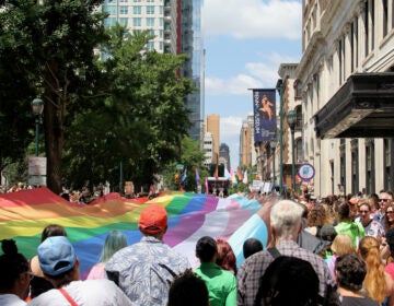 A large LGBTQ flag is carried through the streets by many people.