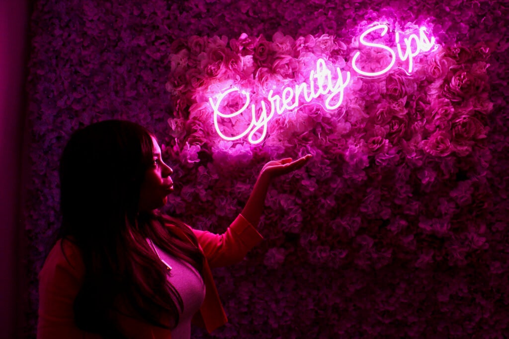 Shakia Williams gestures towards a glowing pink neon sign that reads "Cyrenity Sips"