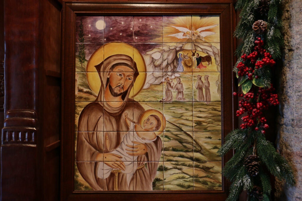 A painted tile mosaic of St. Francis of Assisi holding the baby Jesus and a nativity scene behind him in the distance.