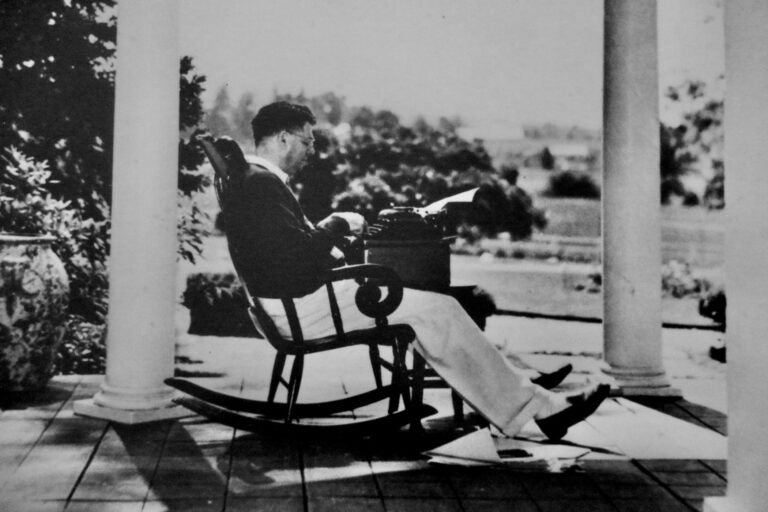 A photo of Oscar Hammerstein writing on a typewriter, seated on a rocking chair on a porch.