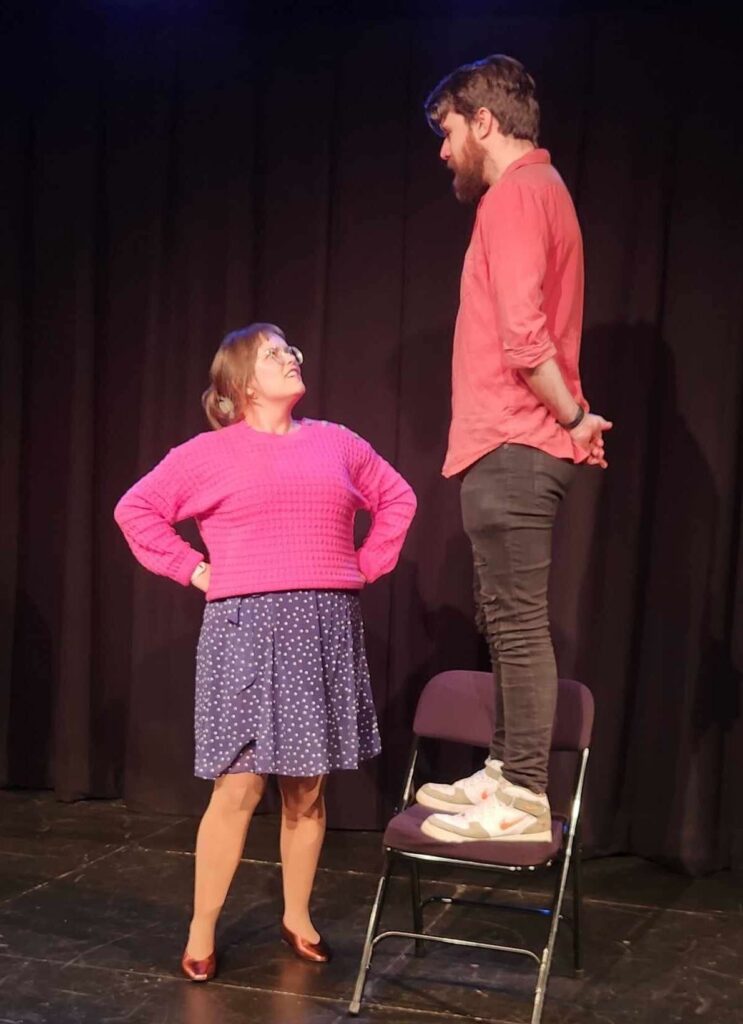 Cara Hammer onstage at an improv group looking up at Jim O'Donnell standing on a chair.
