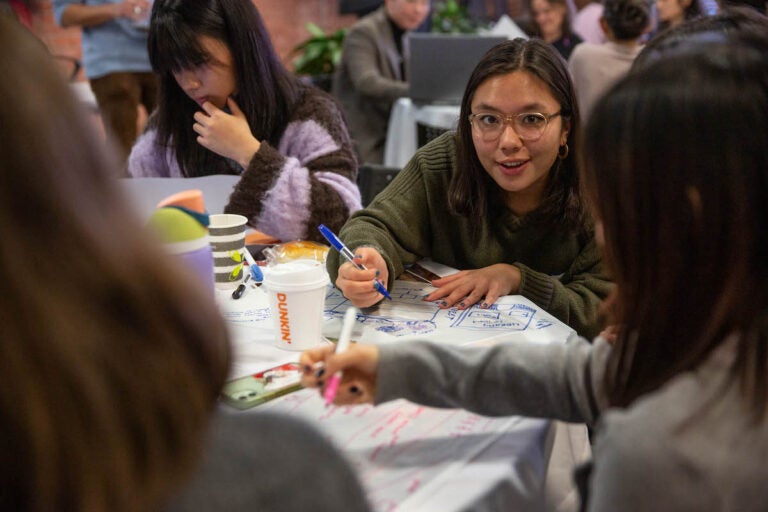 People speak together at a table as they write and draw designs.