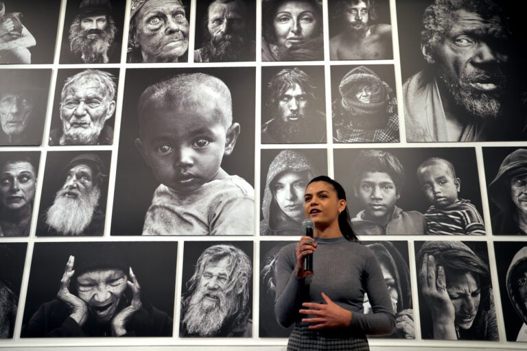 Leah den Bok speaks into a microphone in front of portraits she has made of people experiencing homelessness