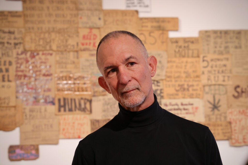 Willie Baronet poses for a photo in front of his work, featuring cardboard signs on the wall.