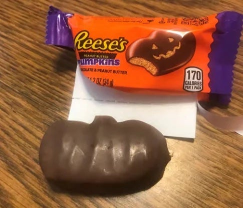 A Reese's Peanut Butter Pumpkin with no carved-out face next to the open wrapper with a jack-o'-lantern design.