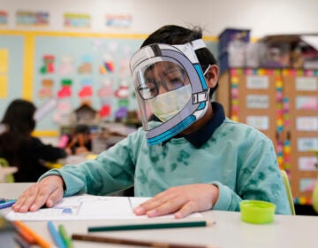 A student wearing a mask and a face shield sits at a desk.