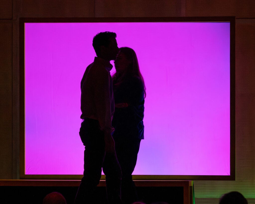 A silhouette of two people embracing onstage in front of a neon pink background