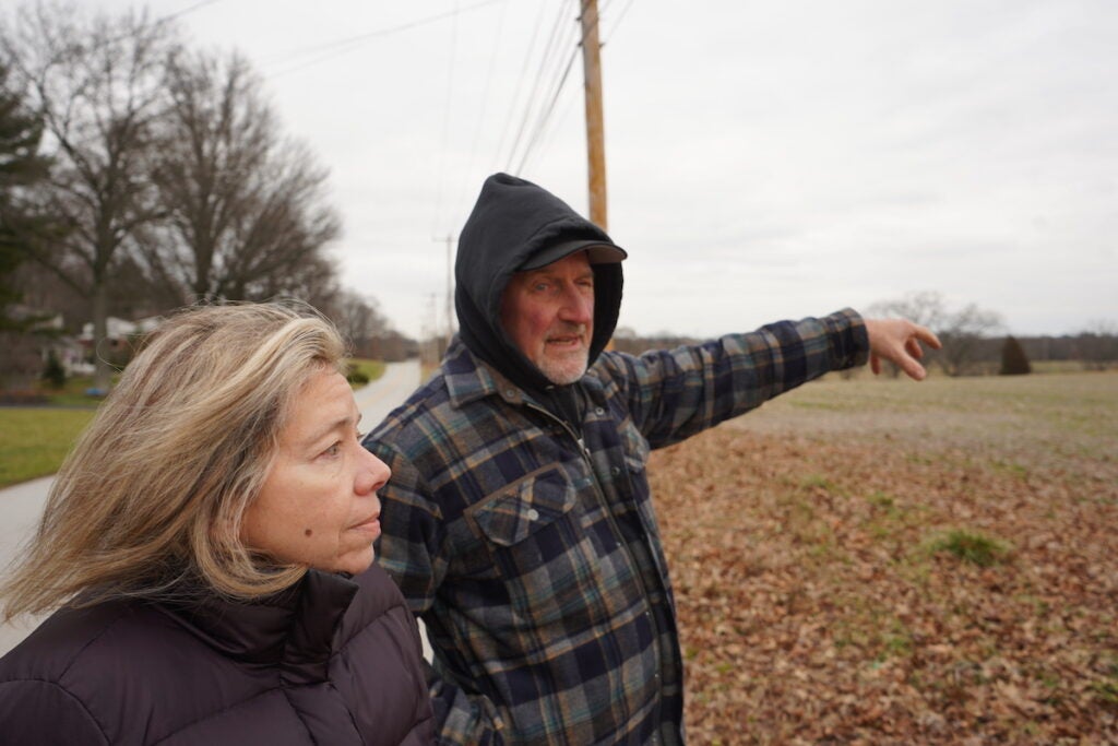Susan Denby, 61, and Bill Felton, 70, stand in front of an open field on a narrow paved road.