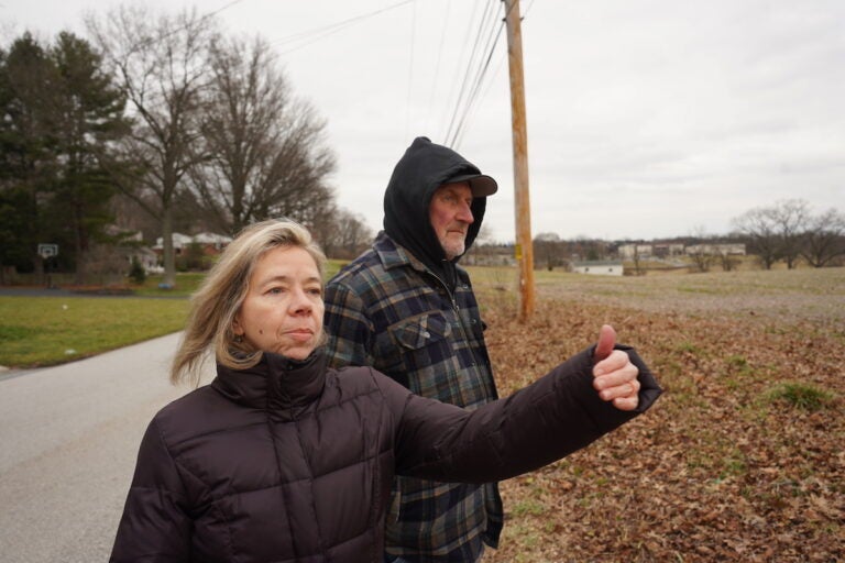Susan Denby, 61, and Bill Felton, 70, stand in front of an open field on a narrow, paved road.