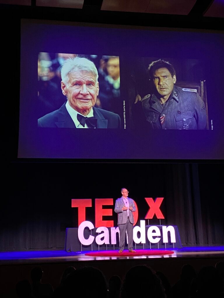 Paul Epstein speaks onstage in front of a slideshow with photos of Harrison Ford