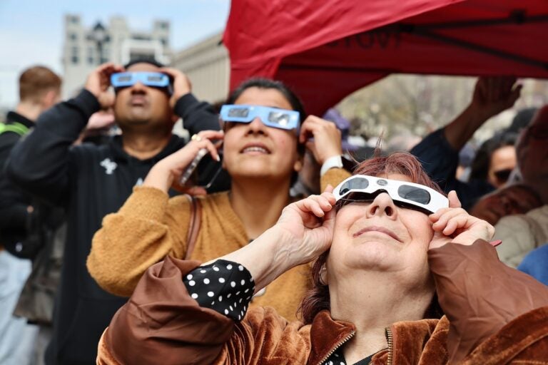 Three people are seen holding up eclipse glasses as the eclipse streaks across Philadelphia