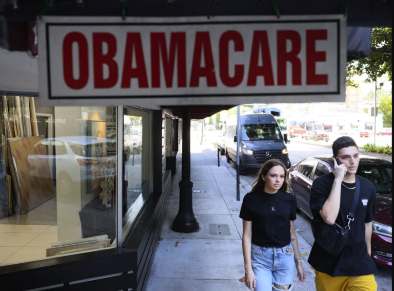 2 people walking on the street passed a sign that says 'Obamacare'