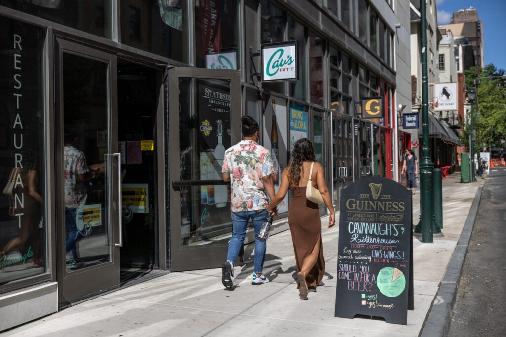 A man and a woman walk hand in hand on the sidewalk past a sign promoting a local restaurant